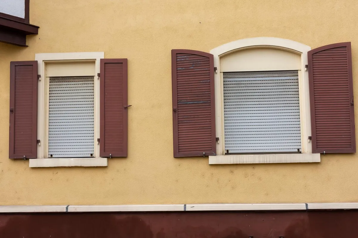 Façade en enduit jaune où des volets roulants clairs côtoient des volets battants en bois brun